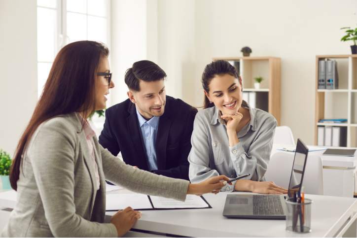 Finance broker meeting with a couple at a desk, reviewing loan documents on a laptop in a bright, neutral office.
