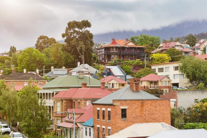 Residential hillside neighbourhood with older-style houses and red roofs among green trees, with a misty mountain range in the background under a cloudy sky.